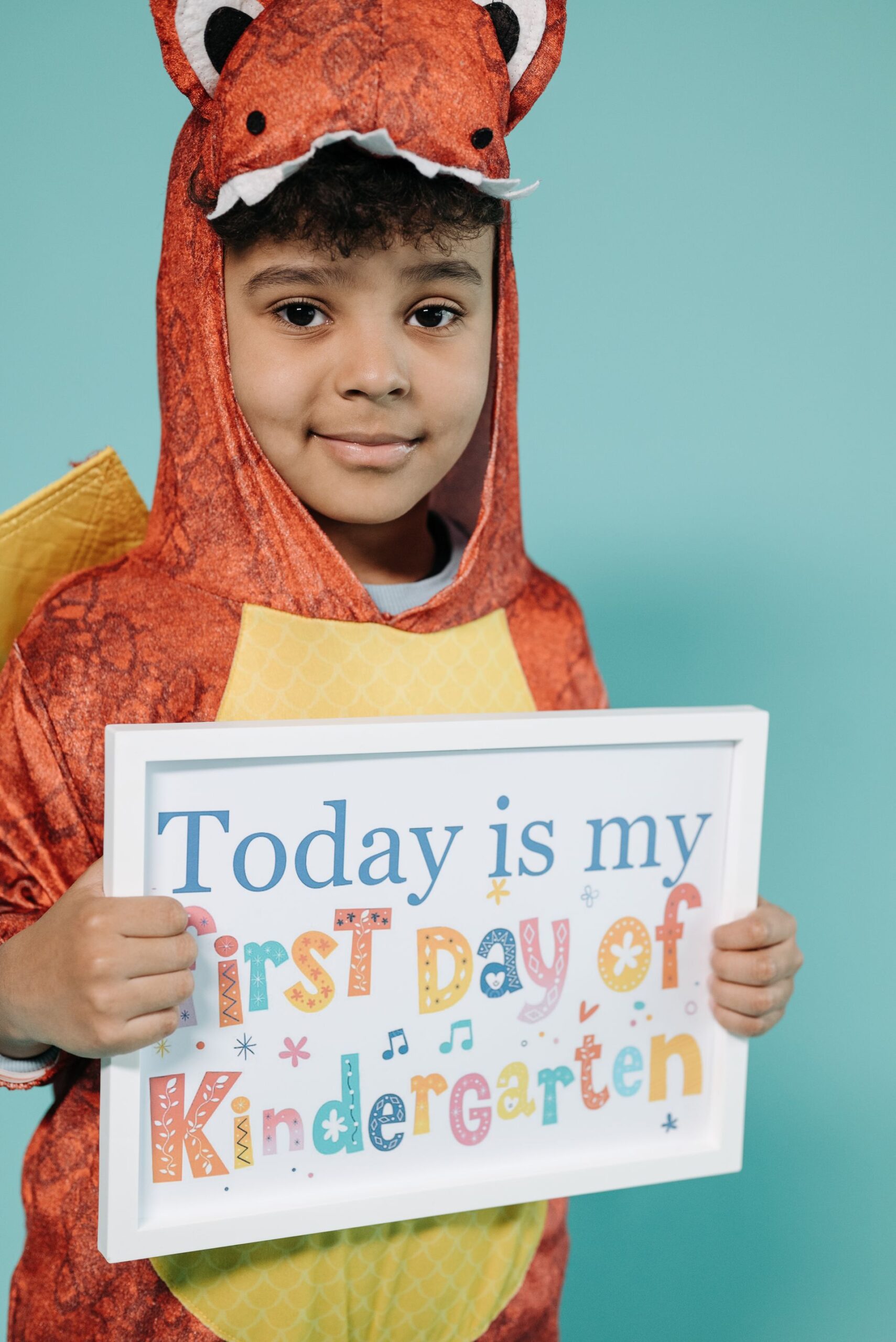 A kindergartener smiling confidently in a dinosaur onesie and holding a “today is my first day of kindergarten” sign. Rawr, it’s a good outfit idea for kindergarten orientation day.